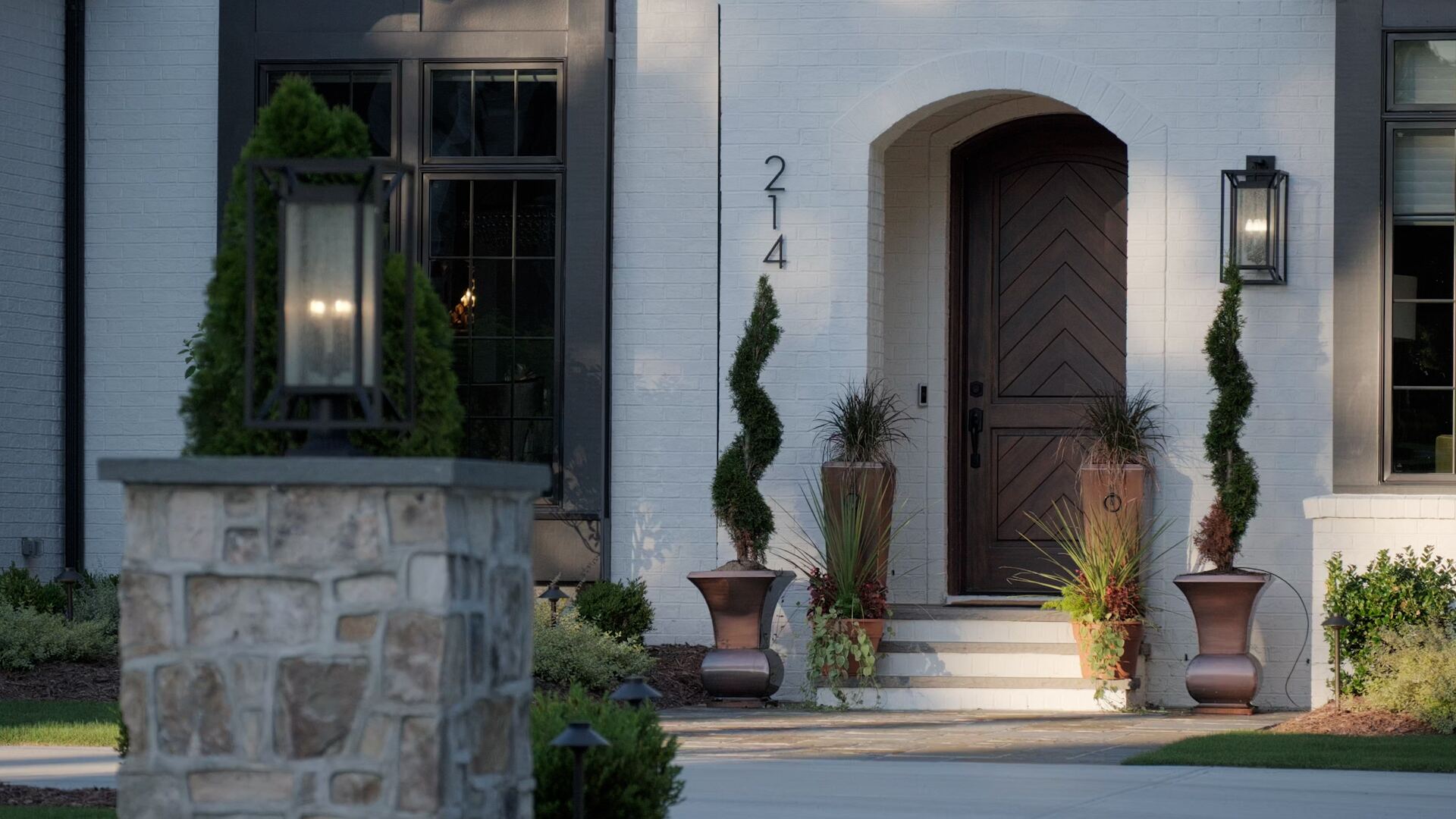 The arched wood front door of a white brick Montlor home at dusk, framed by lanterns and topiaries.